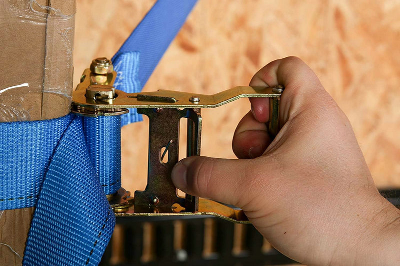 A blue 1.5-inch ratchet strap (LC 1250kg) is tightened around a box with a ratchet buckle.