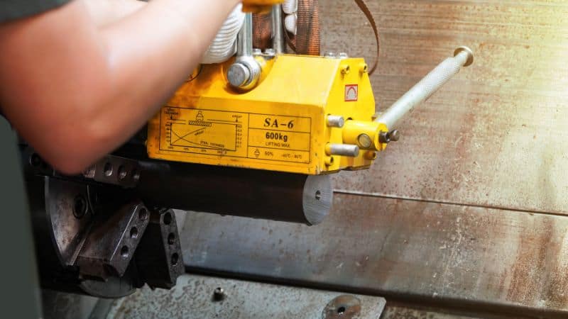 Worker lifts a cylindrical metal piece with a yellow SA-6 600 kg magnetic lifter on a workshop floor.