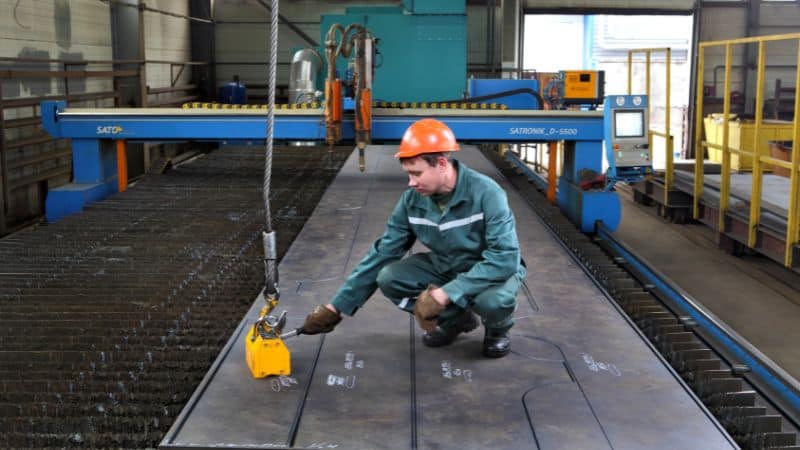 A worker in safety gear uses a magnetic lifter to handle a large metal sheet in an industrial facility.